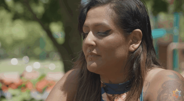 A person with long brown hair and visible piercings sits outdoors, eyes closed, with blurred greenery and colorful objects in the background.
